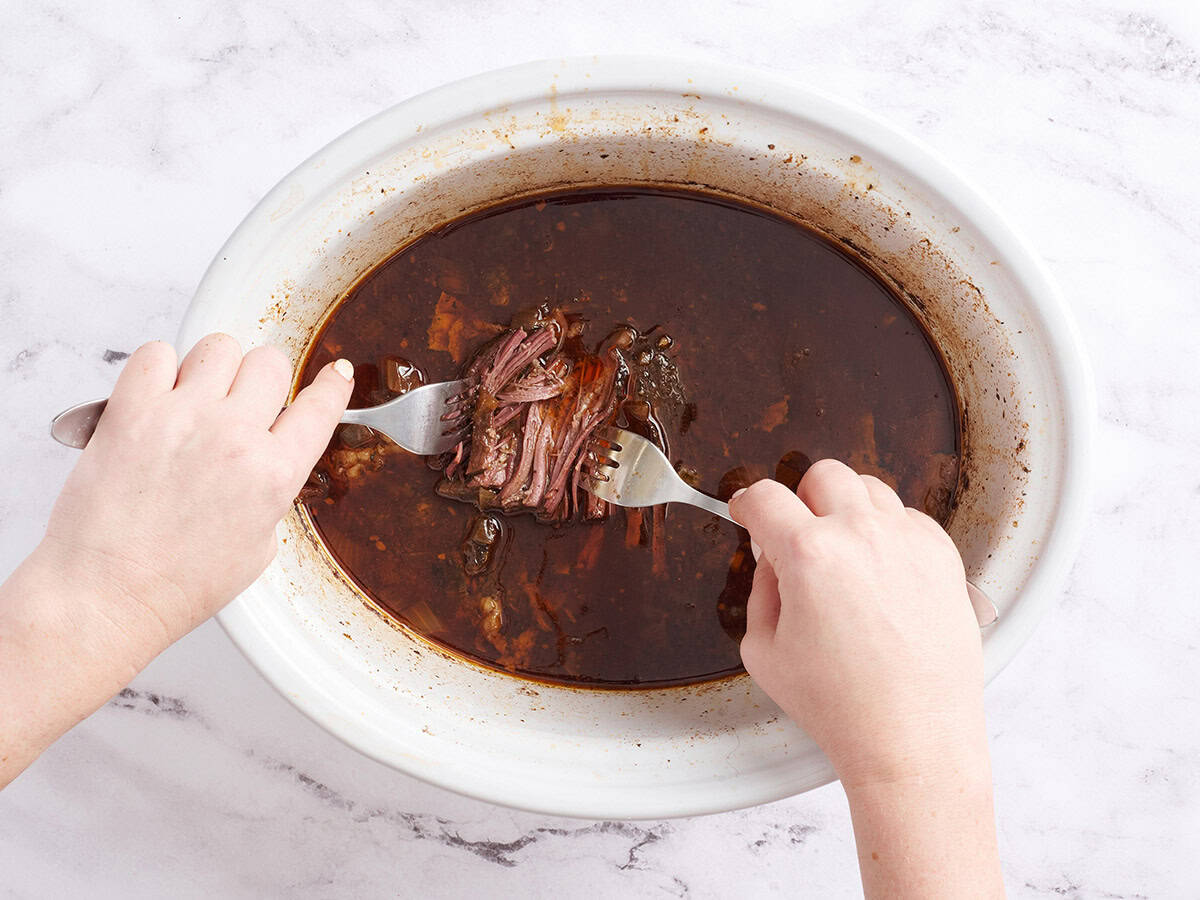 Hands using two forks to shred the meat for beef birria tacos in a crockpot.