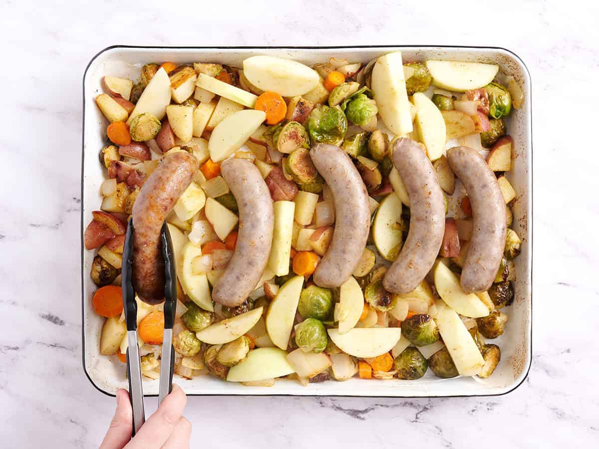 Kitchen tongs flipping a bratwurst sausage over on a sheet pan.