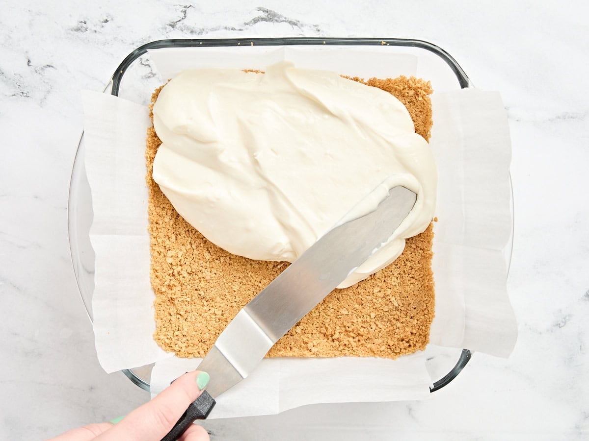 A palate knife spreading a cheesecake filling over a graham cracker crust.