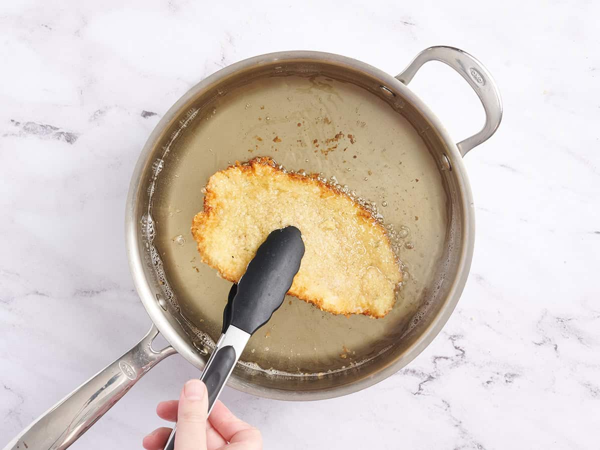Chicken tongs flipping a breaded chicken schnitzel in hot oil.