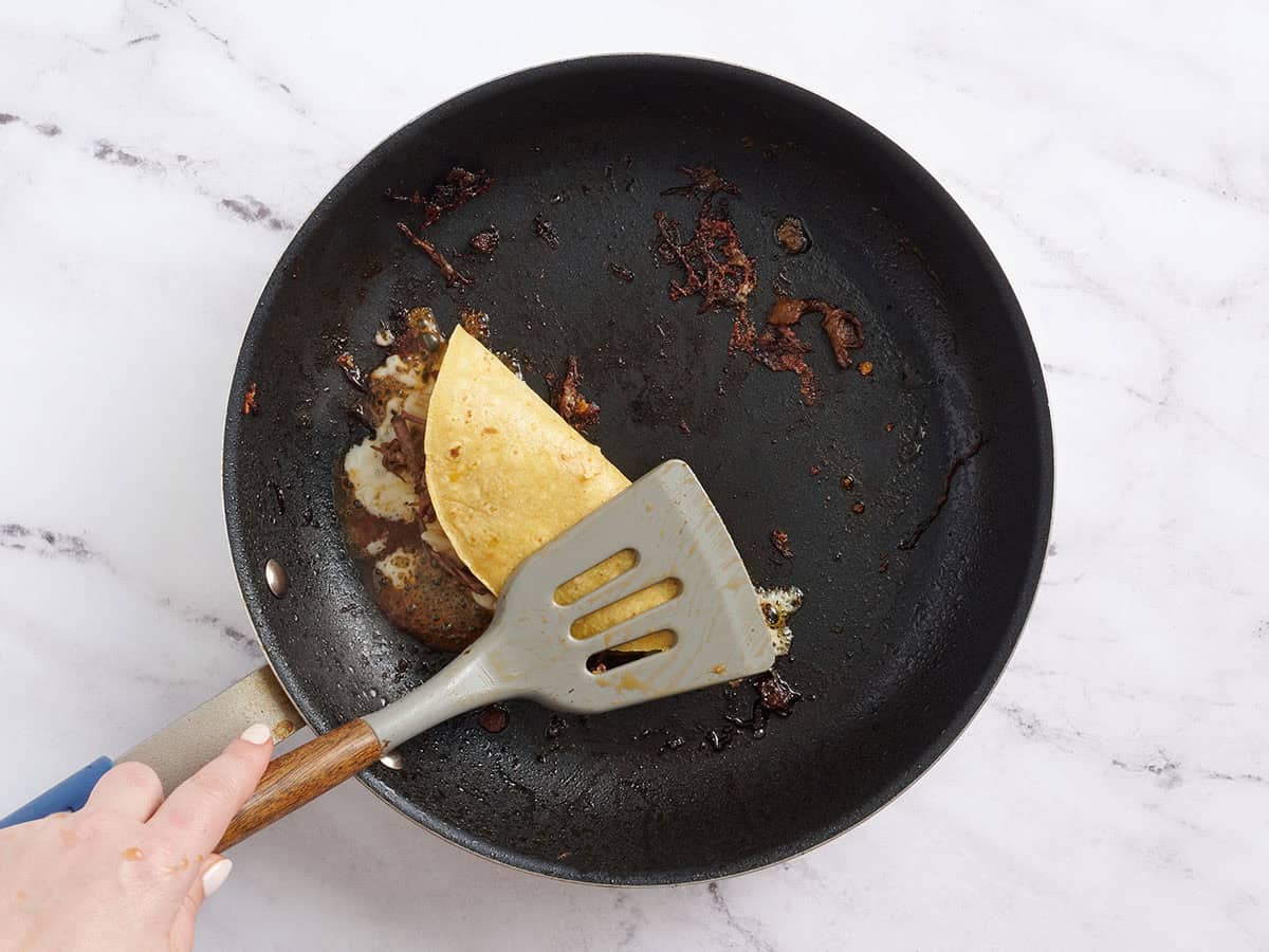 A spatula pressing down a birria taco in a skillet.