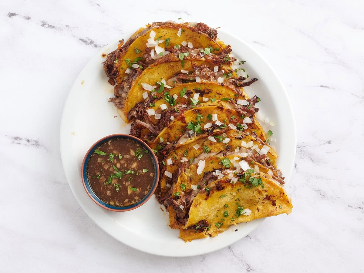 A platter of homemade birria tacos with a pot of beef broth for dipping.