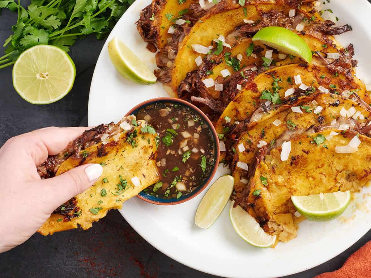 Overhead view of birria tacos on a plate with a hand dipping one into a pot of beef consome.