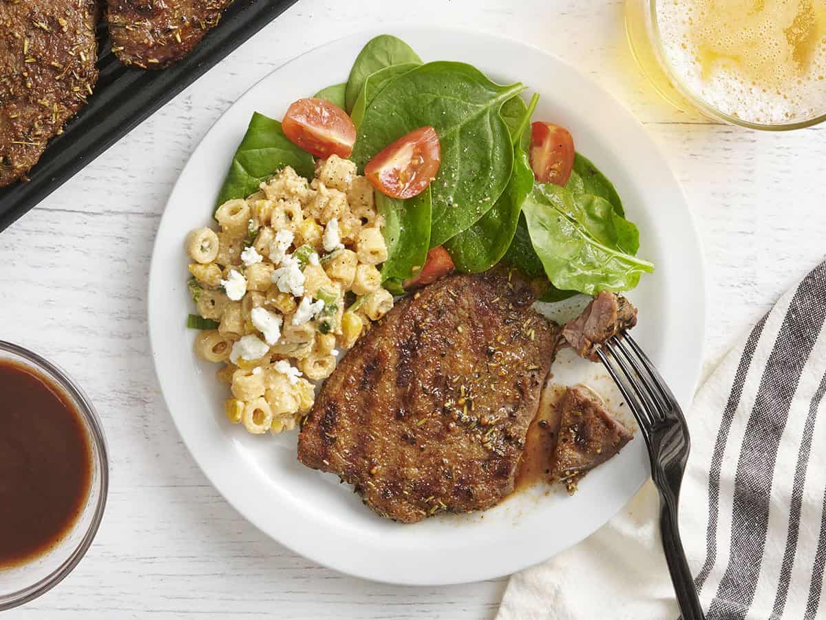 Overhead view of a grilled sirloin tender steak on a plate with pasta salad, spinach, and tomatoes, with a fork taking some.