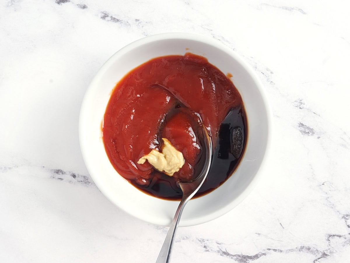 Dijon mustard, ketchup, soy sauce, and brown sugar being mixed together with a spoon in a bowl.