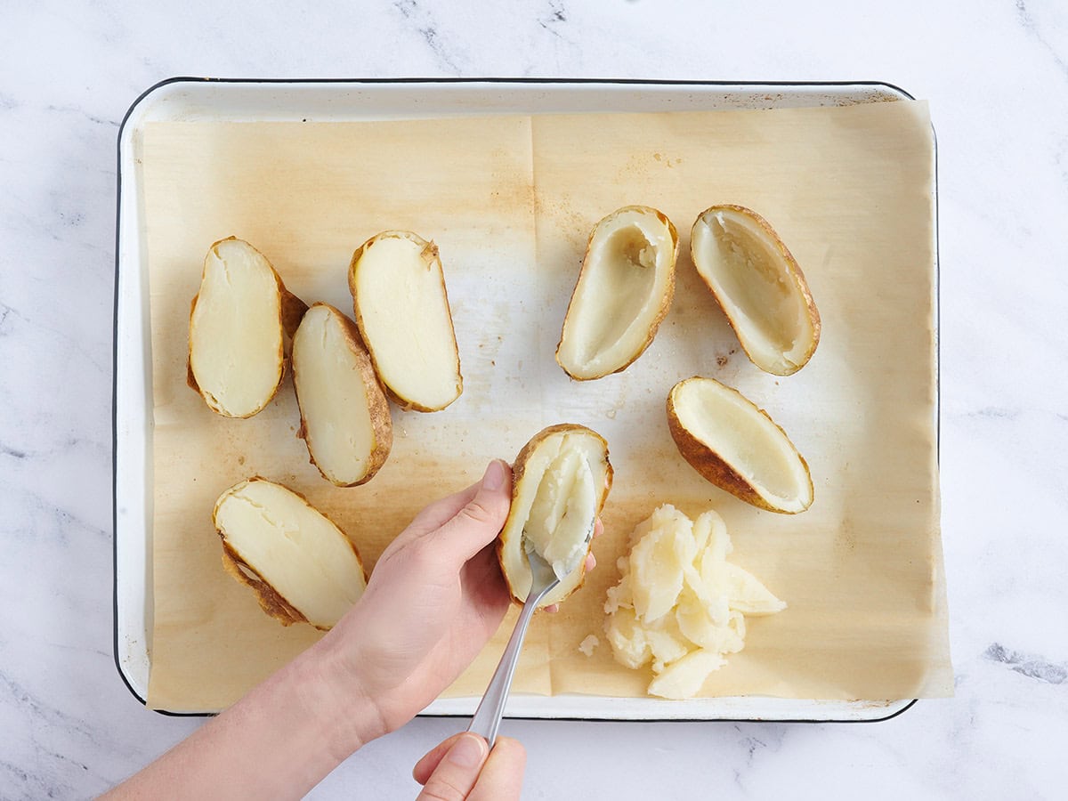 Hands using a spoon to scoop the flesh from baked potato halves.