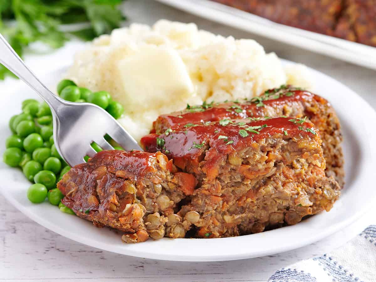 A slice of lentil loaf on a plate with peas and mashed potato with a fork taking some.