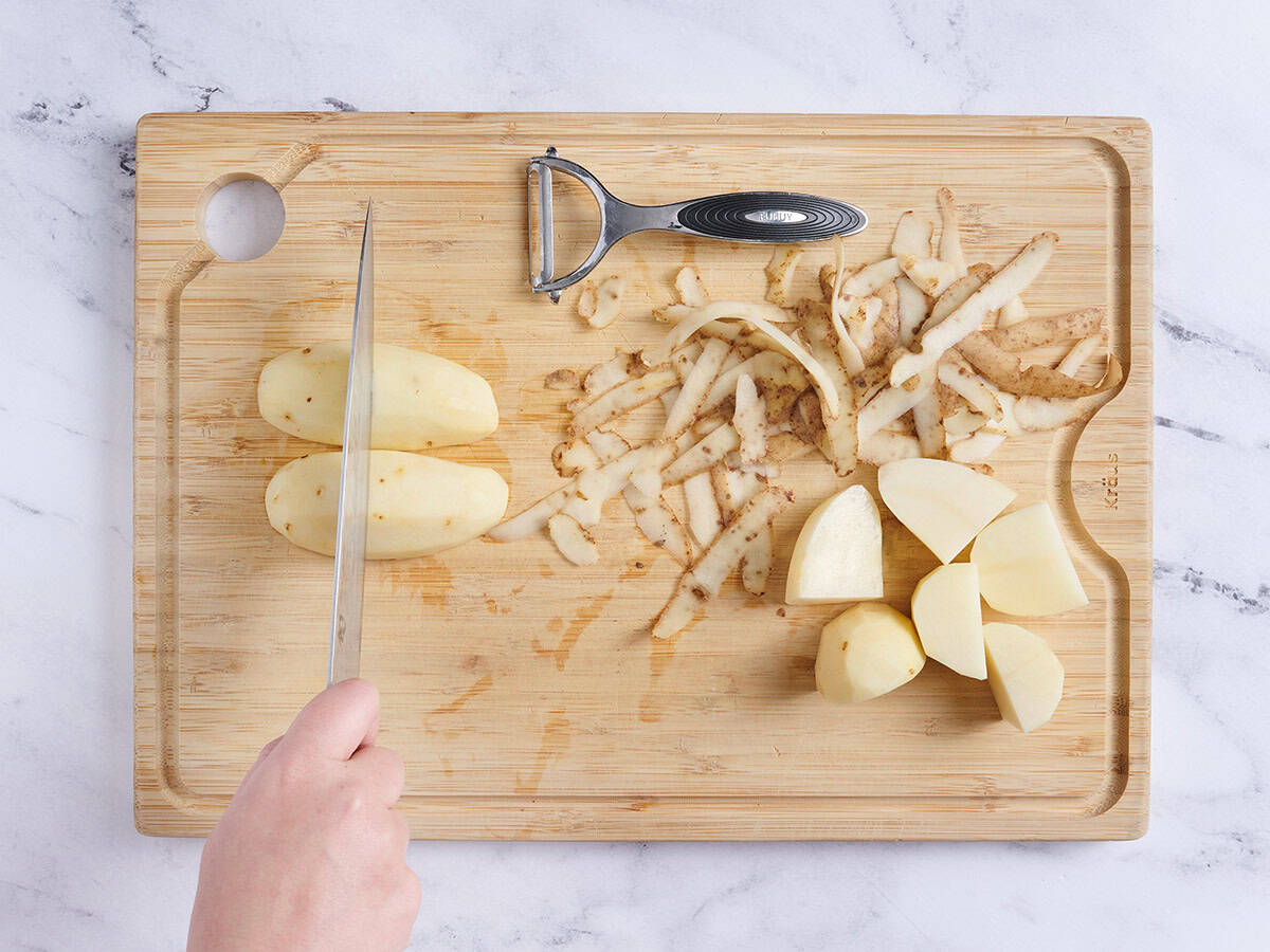 Peeled potatoes on a wooden cutting board being sliced with a knife.
