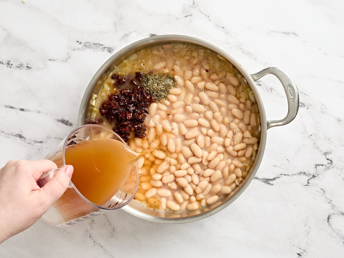 Spices and broth added to white beans in a skillet.