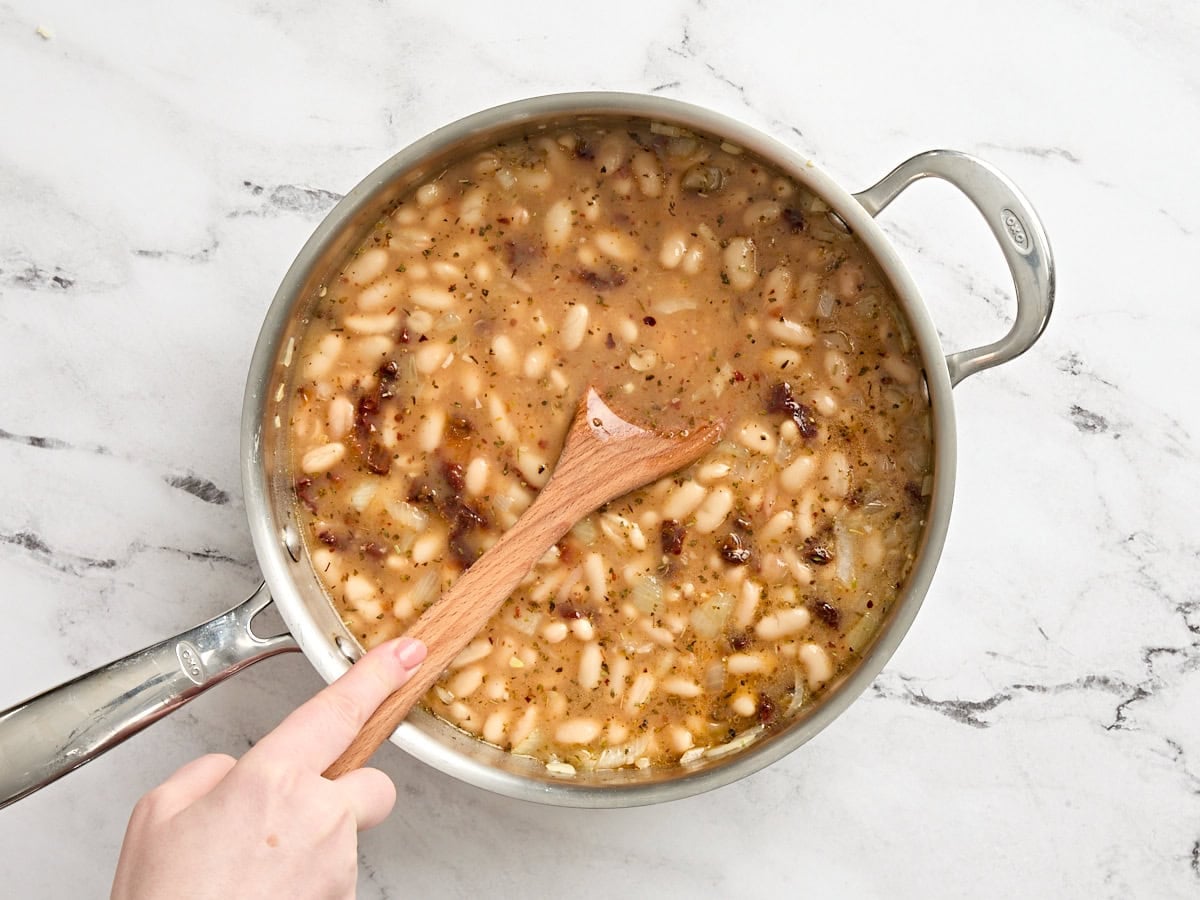 A wooden spoon mashing some white beans in a skillet.