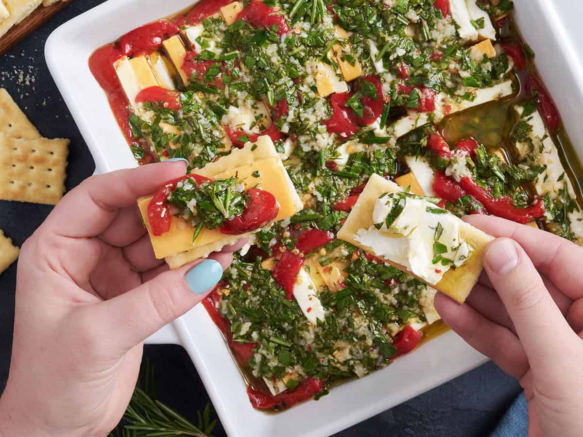 Overhead view of a pan of marinated cheese, with two hands holding a cracker with cheese on.