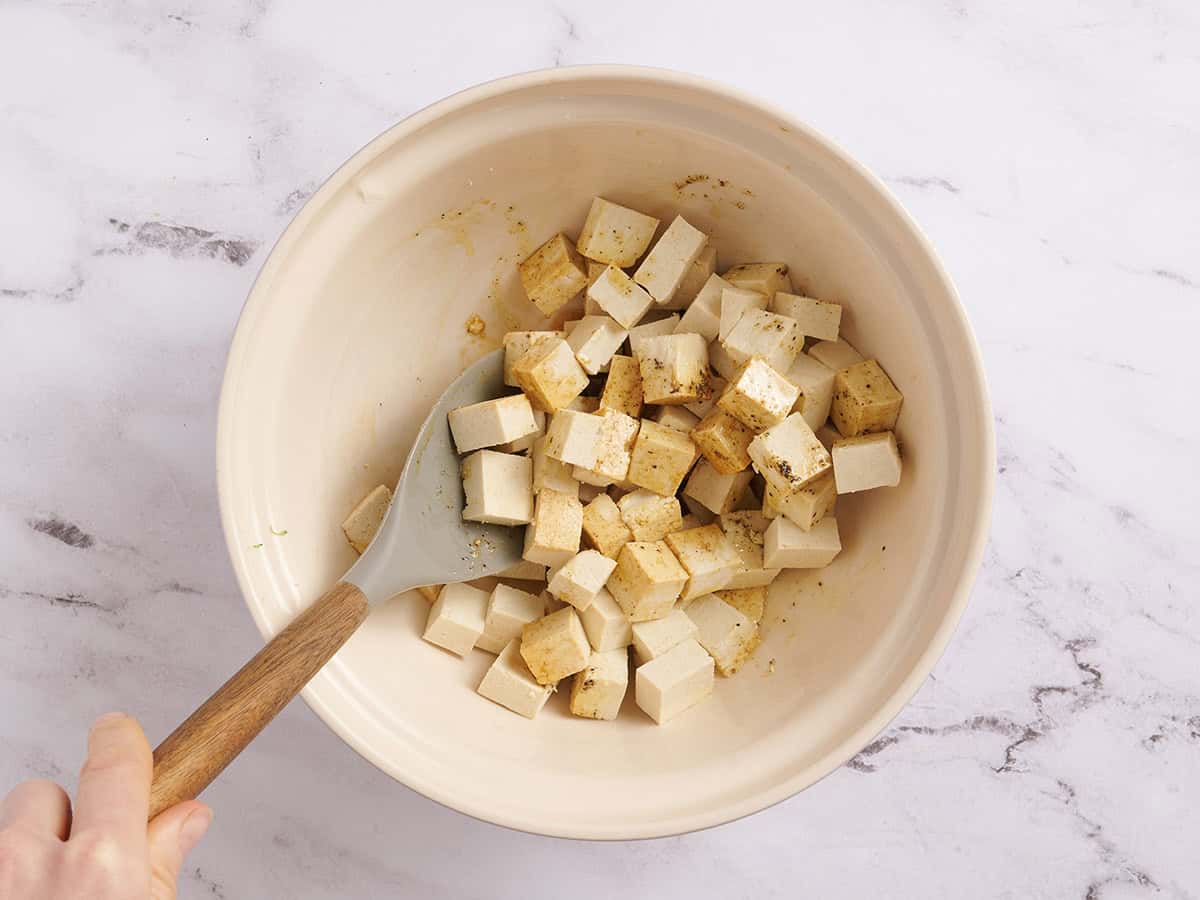 Tofu cubes being tossed with oil, soy sauce and seasonings in a bowl with a wooden spoon.