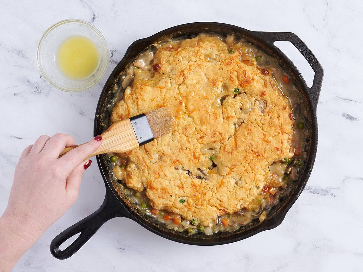 A hand brushing melted butter on a chicken cobbler.