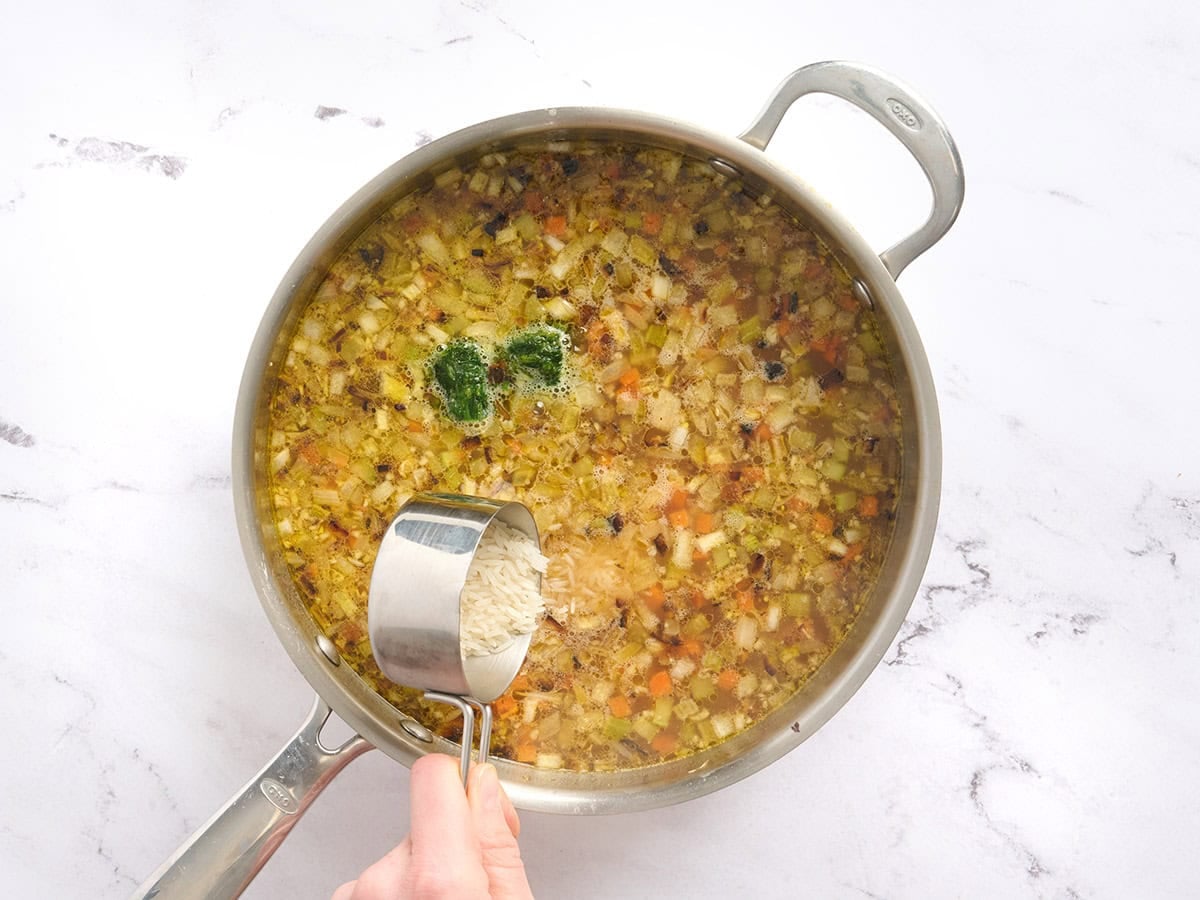 Rice being added to a skillet of chicken broth soup.