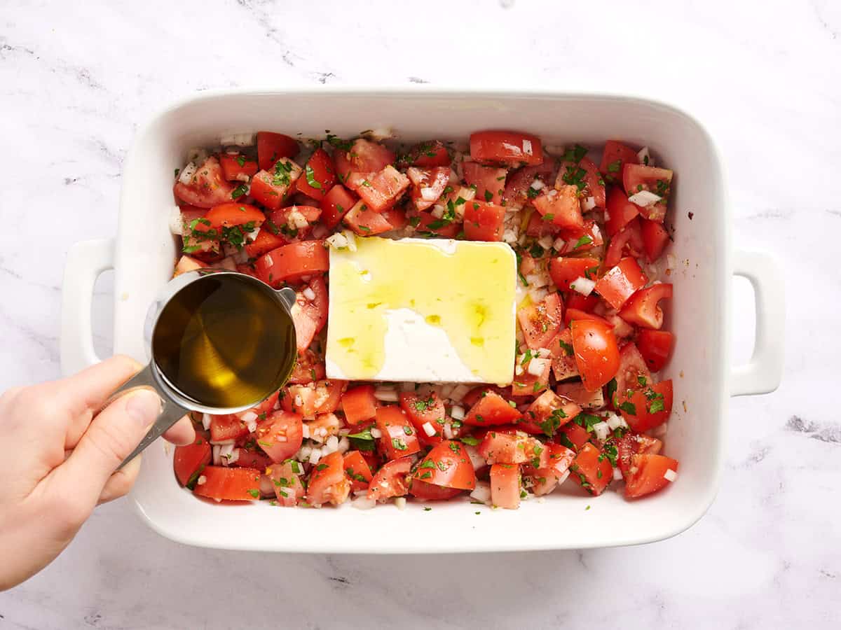 A hand drizzling olive oil over tomatoes, onions, and feta cheese.