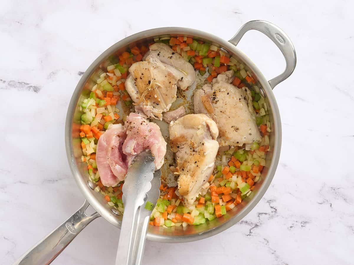 Chicken thighs being cooked in a skillet with mirepoix.