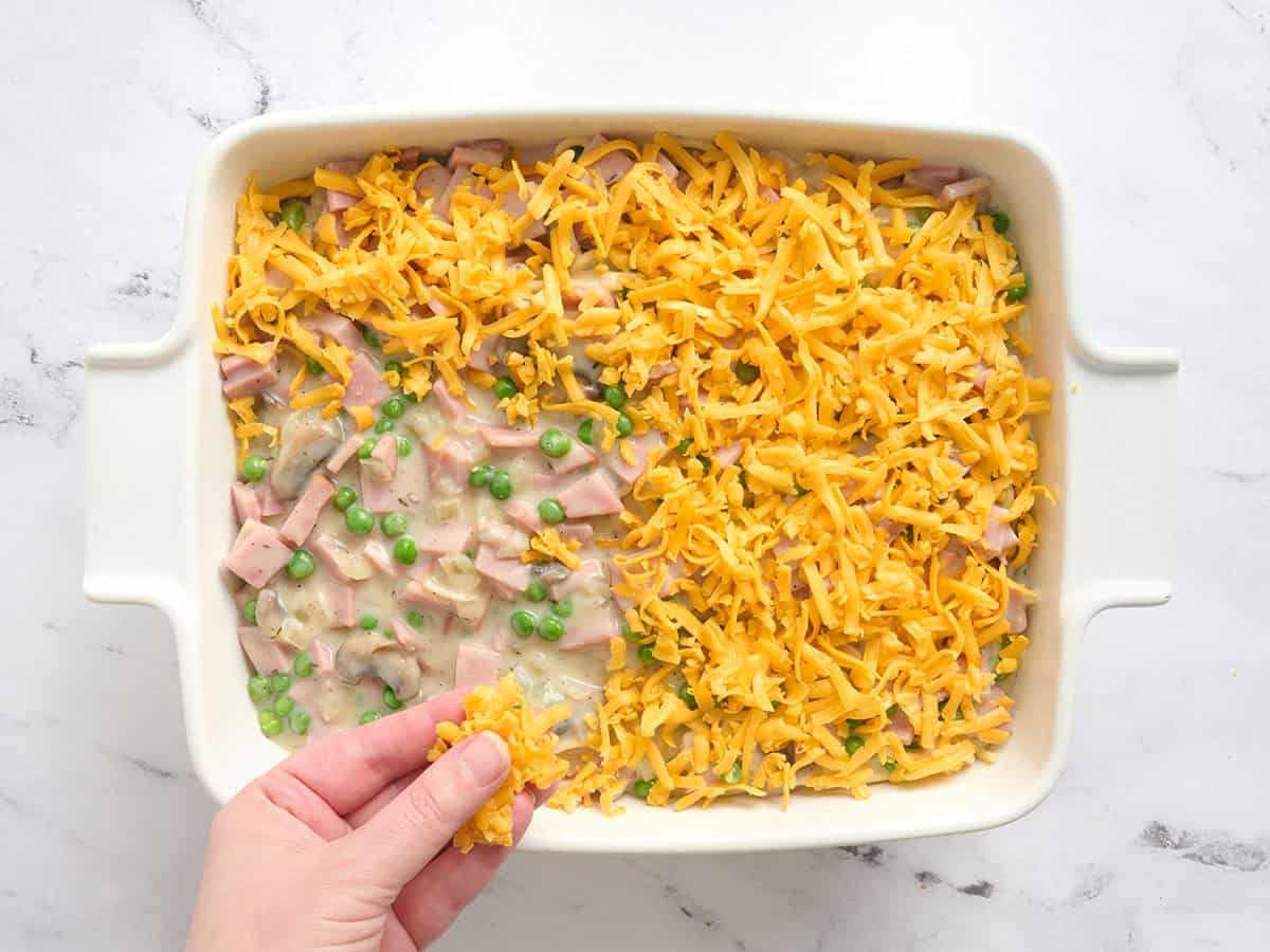 A hand sprinkling cheddar on a ham tetrazzini.