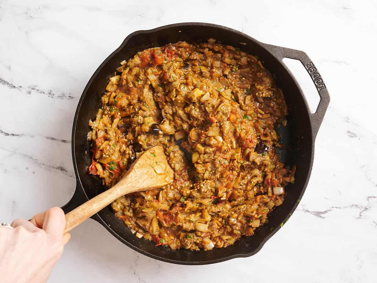 A wooden spoon mashing eggplant for a curry in a skillet.