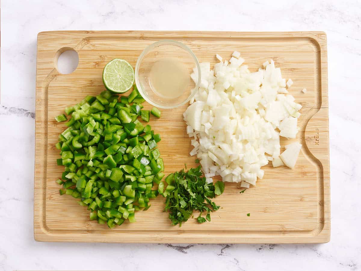 Diced green bell pepper, diced onion, chopped cilantro, and lime on a wooden cutting board.