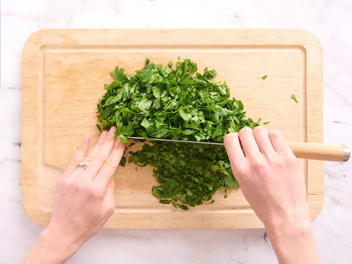 Hands using a knife to mince herbs on a wooden cutting board.