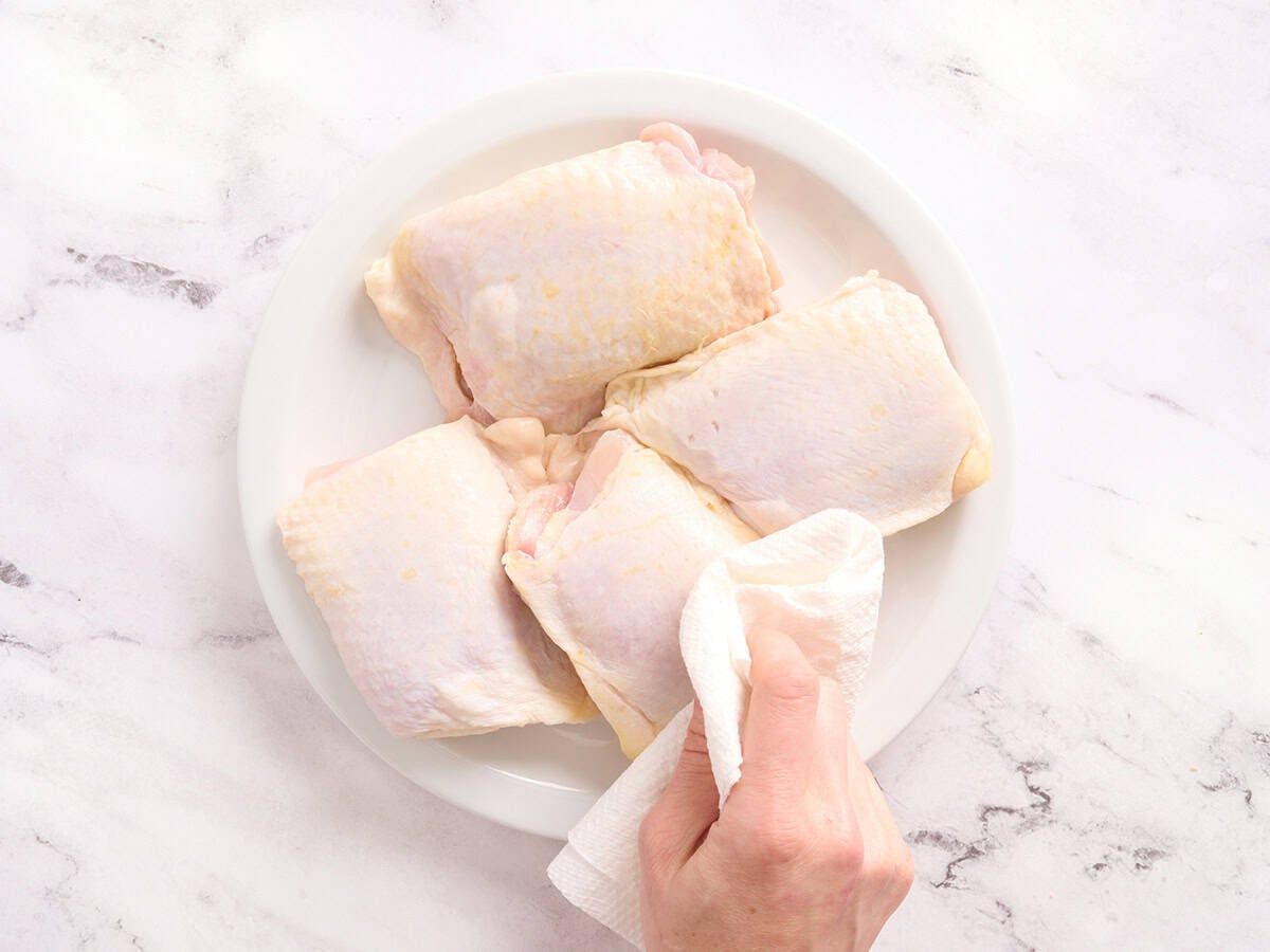 A hand using a paper towel to pat chicken thighs dry.