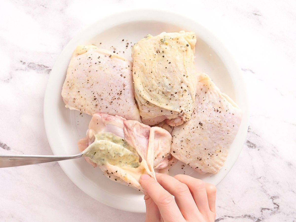 Hands pulling back the skin on a chicken thigh and adding chimichurri marinade.