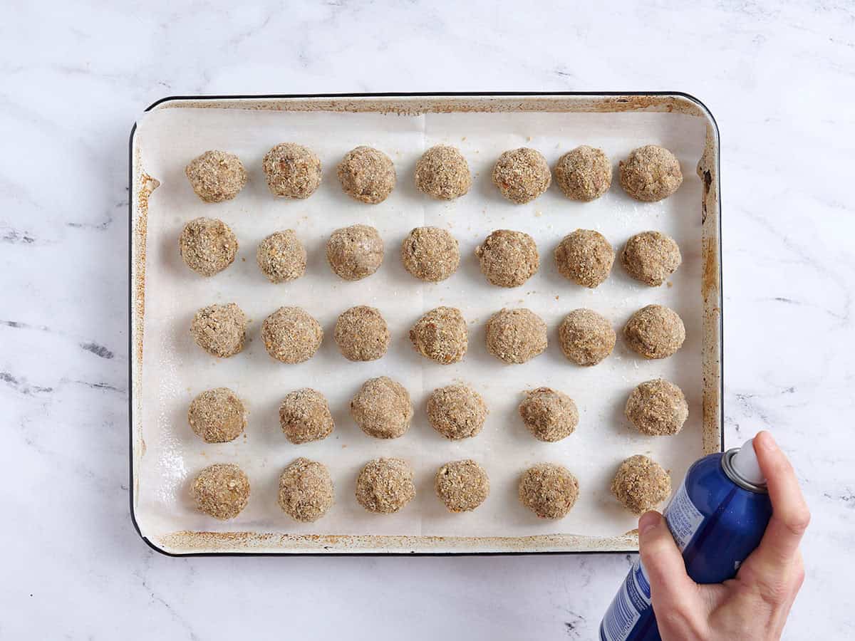 Vegan meatballs on a baking sheet being sprayed with cooking spray.