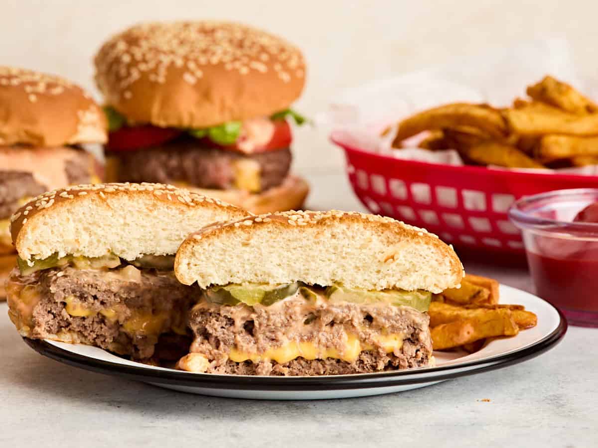 Side view of a juicy lucy burger sliced in half on a plate with more burgers and a basket of fries in the background.