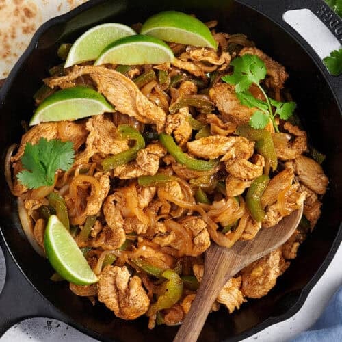 Overhead view of stovetop chicken fajita filling in a skillet with a wooden spoon.