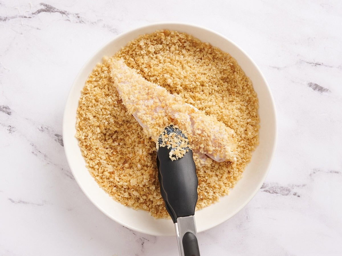 A chicken tender being tossed in toasted breadcrumbs in a bowl.