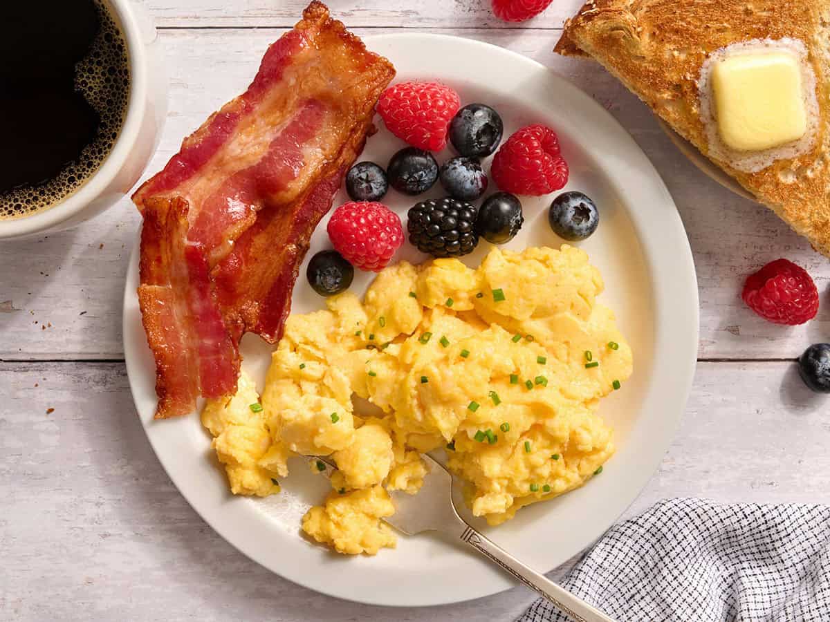 Overhead view of cottage cheese eggs on a plate with bacon and a fruit salad.