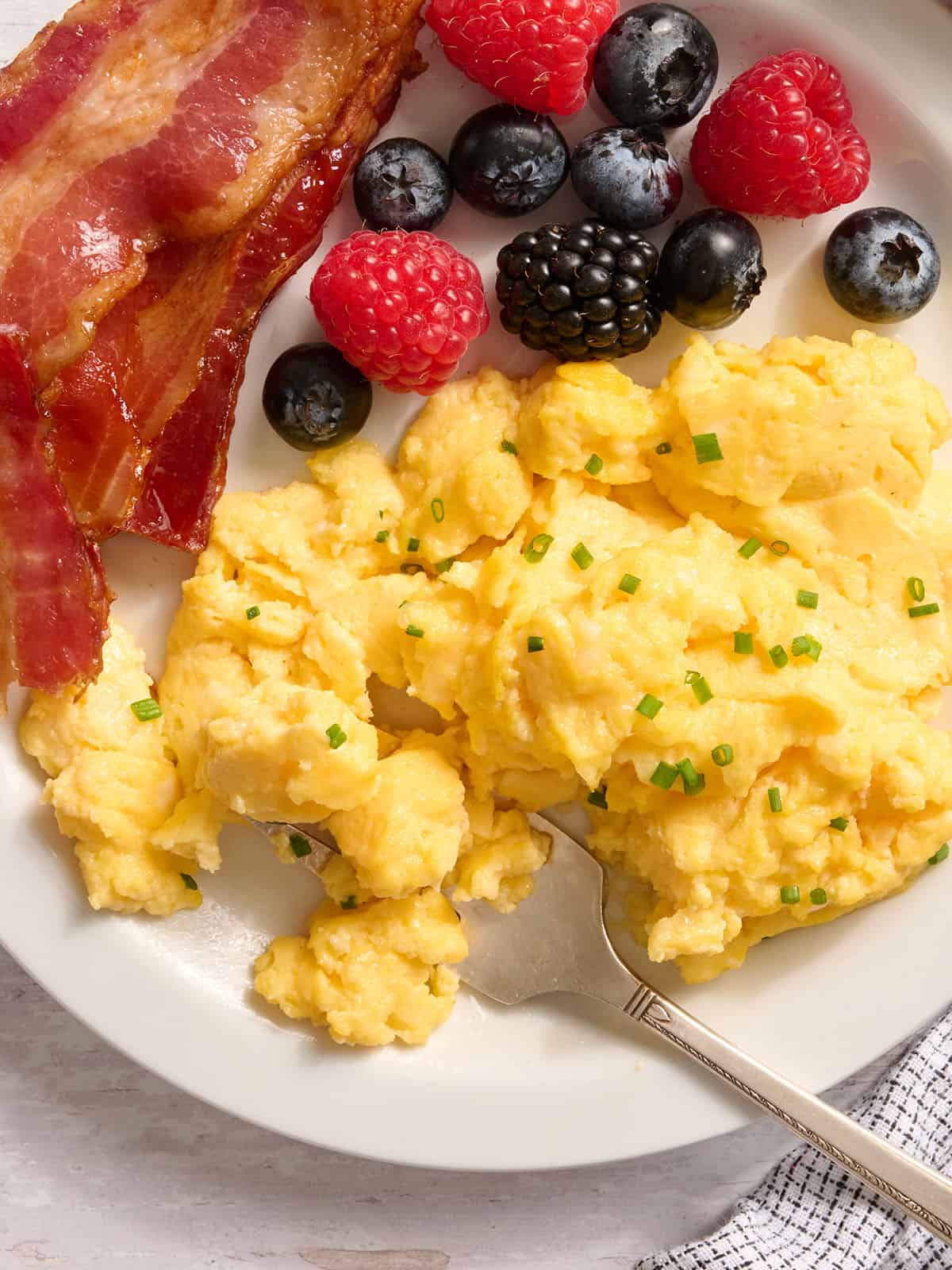 Overhead view of cottage cheese eggs on a plate with bacon and a fruit salad.
